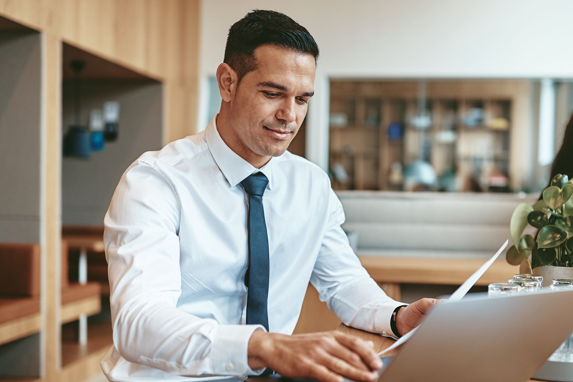 A professional wearing a white shirt and tie sits at a desk, reviewing a document while working on a laptop.