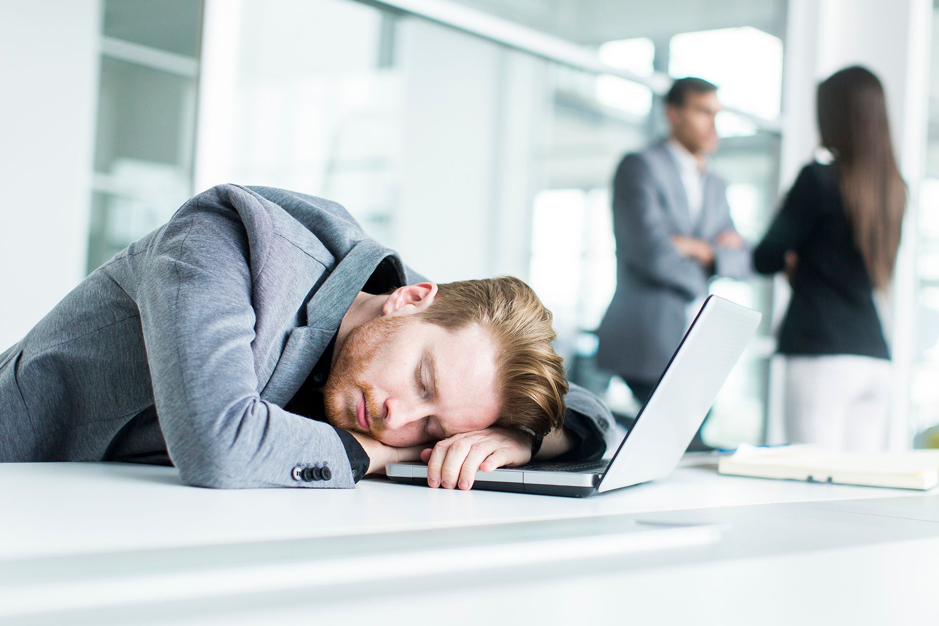 A person in a business suit sleeping with their head on a laptop on an office desk, with two coworkers in the background.