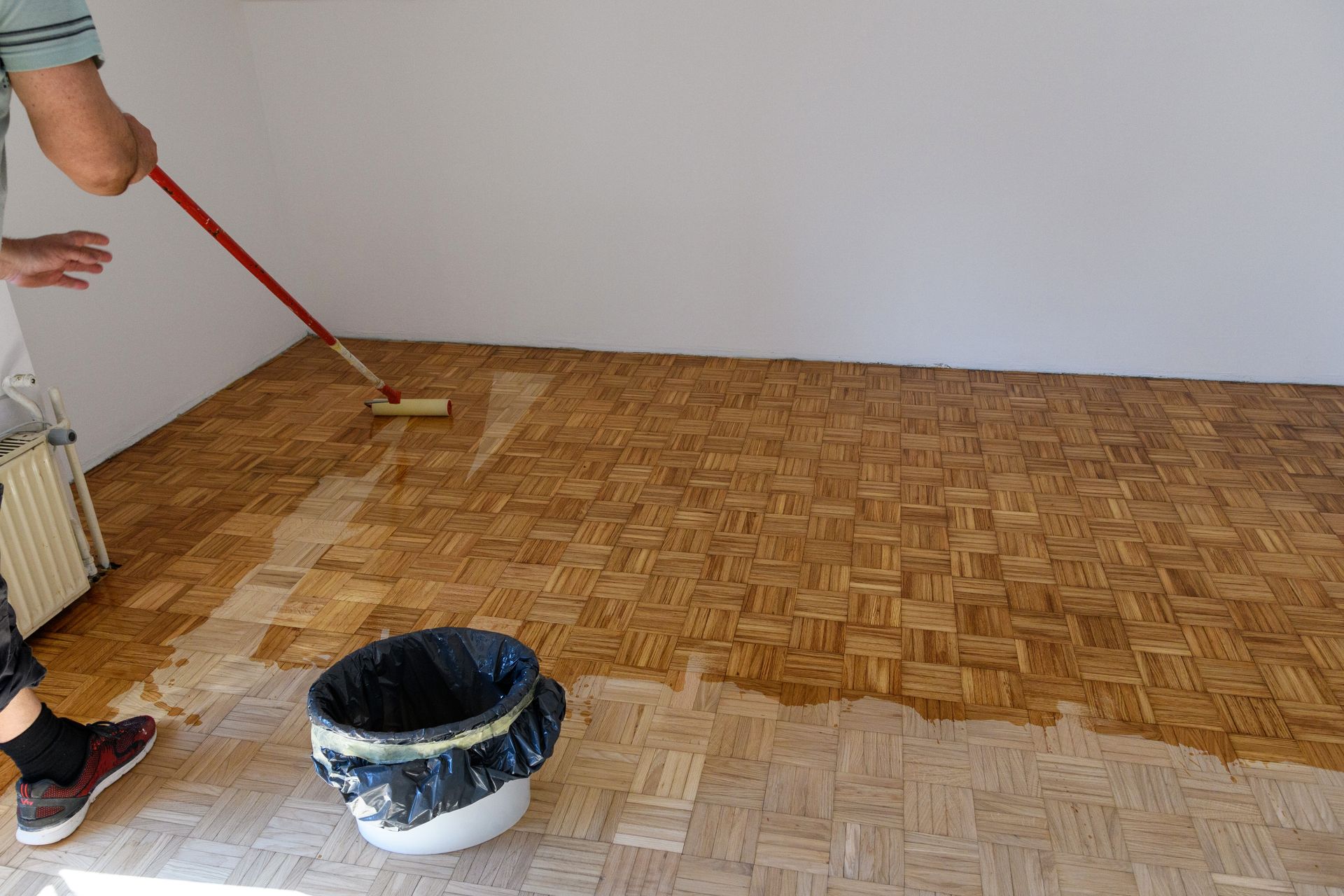 Person painting a parquet floor brown with a roller beside a paint bucket