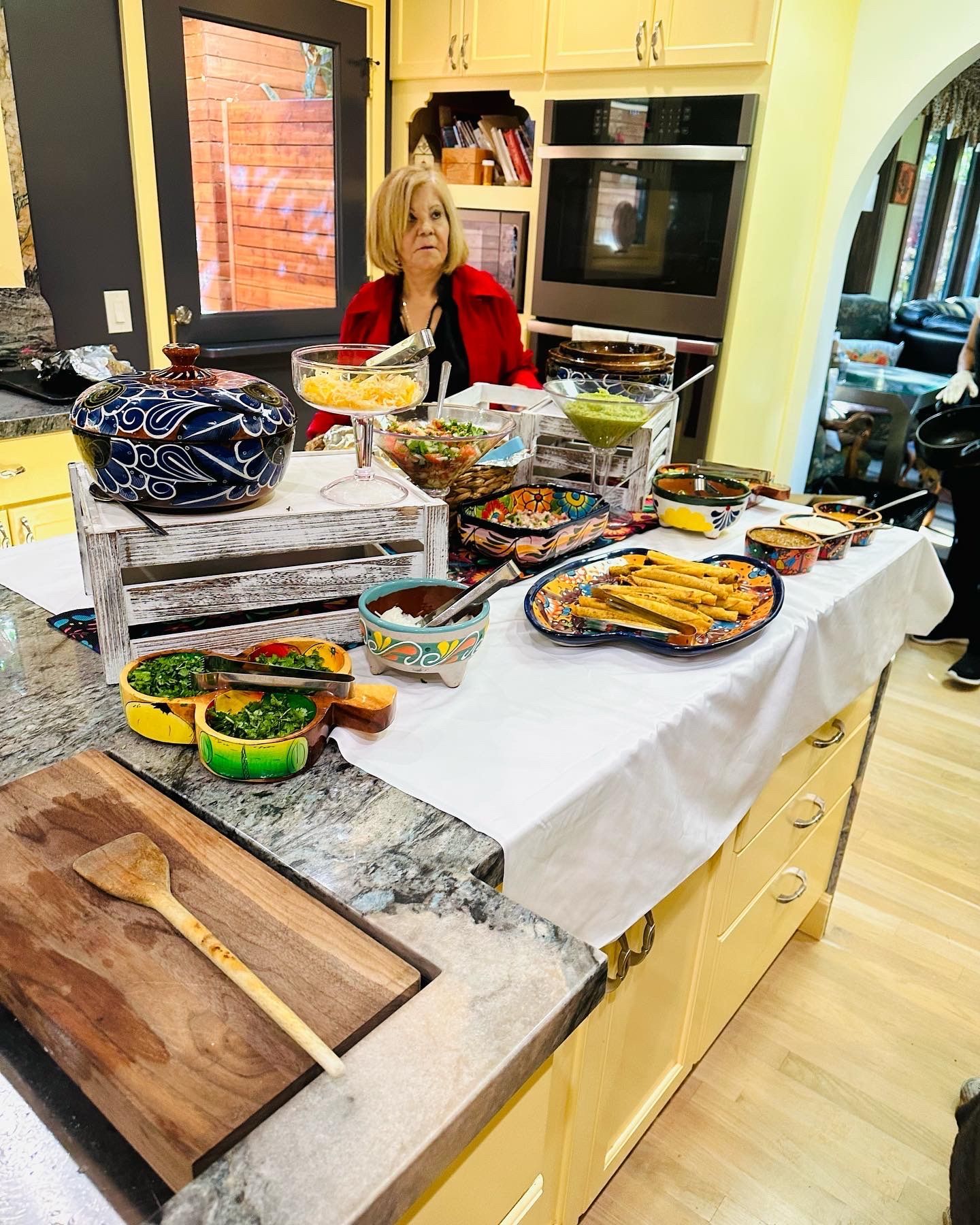 Woman in a kitchen, standing behind a table laden with various dishes and a wooden cutting board.