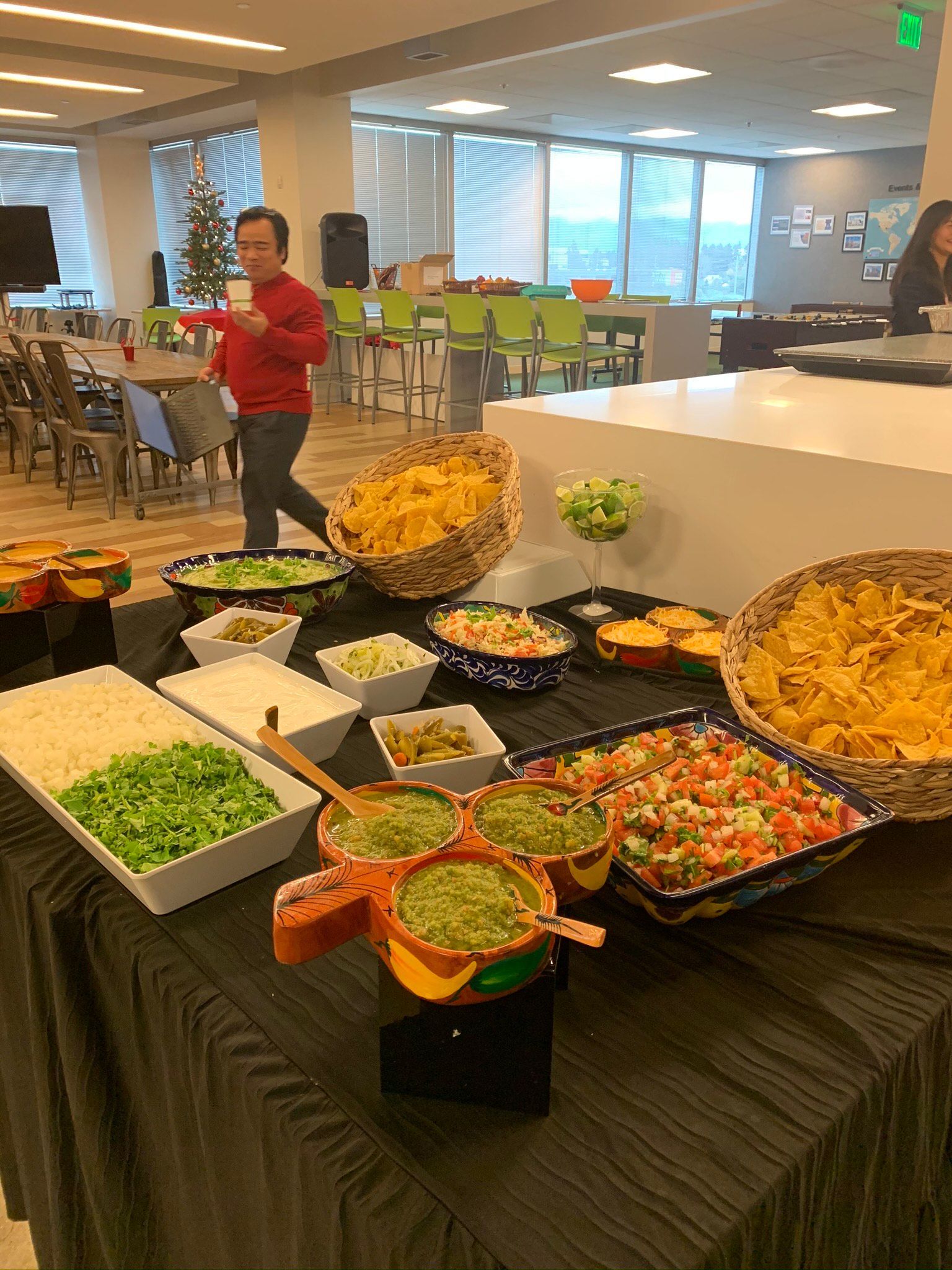 Buffet of taco fixings; person in red shirt walks by. Bright office setting.