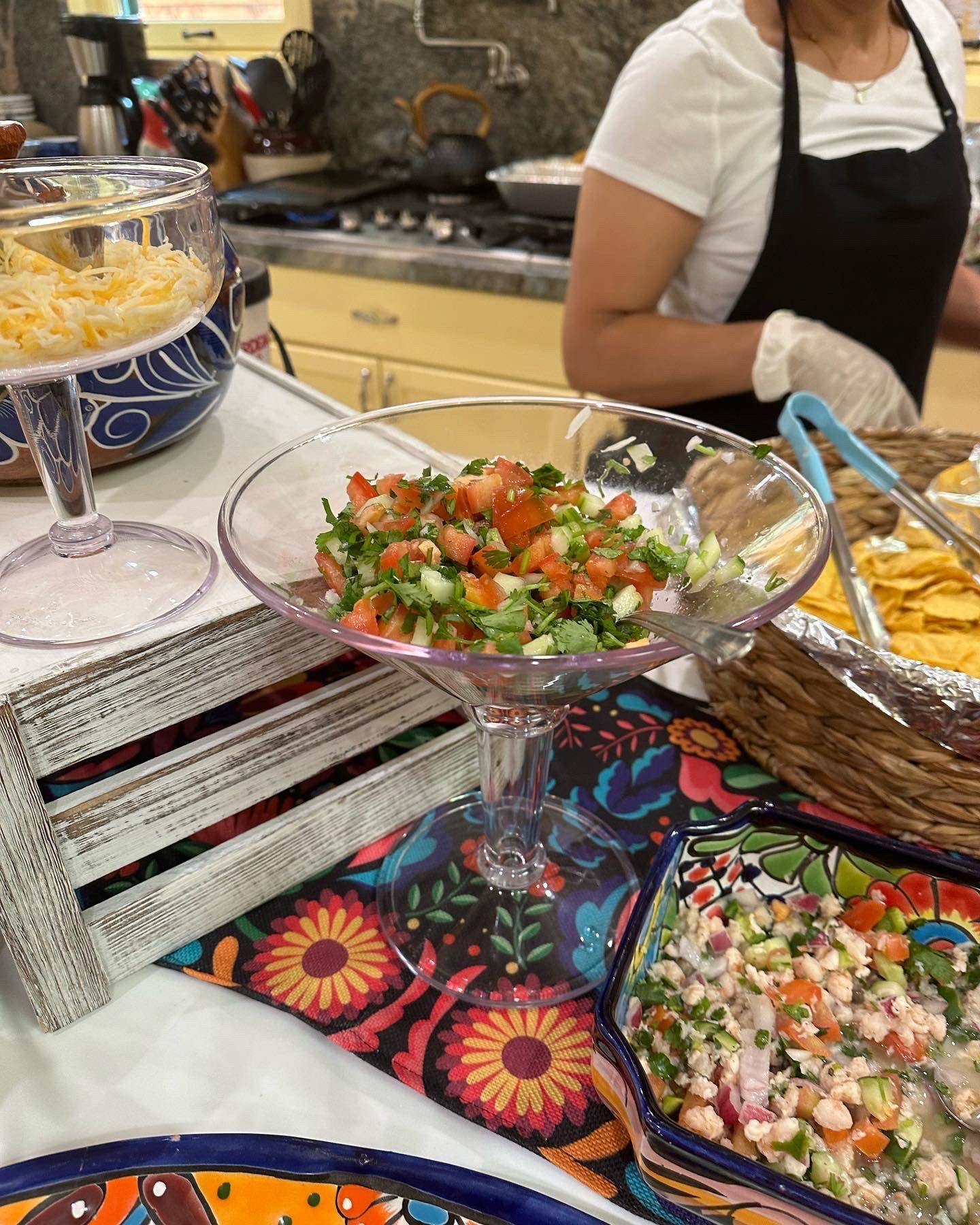 Table with food: Pico de gallo, shredded cheese, chips, and woman with apron.