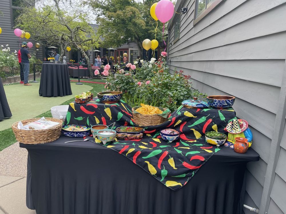 Buffet table with bowls of food on chili pepper-patterned cloth, outside, balloons in background.