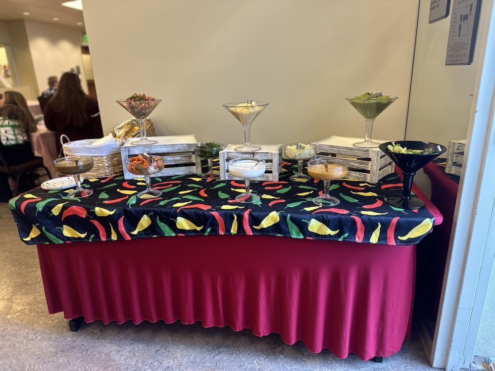 Buffet table with appetizers, red skirt, black tablecloth with chili peppers, white boxes.