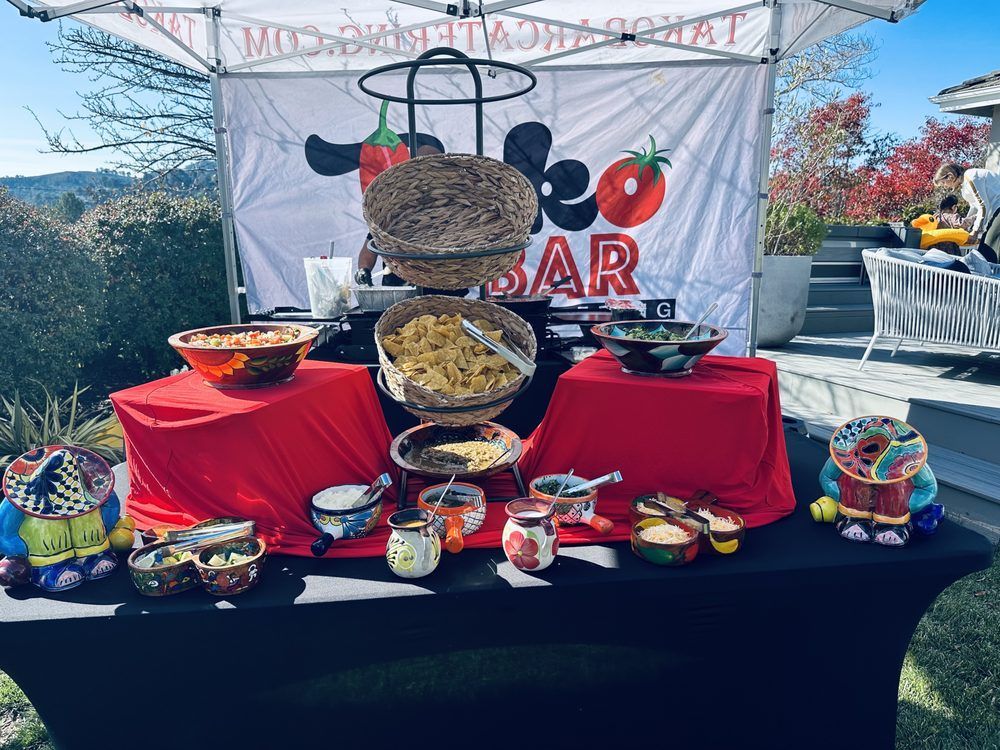 Taco bar setup with colorful food bowls on red and black covered tables, outdoors.