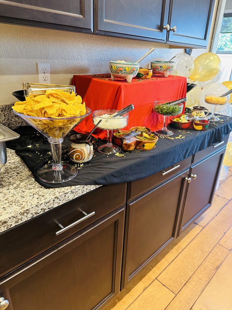 Snack bar with chips, dips, and toppings on a black tablecloth over dark brown cabinets; a red stand holds some bowls.