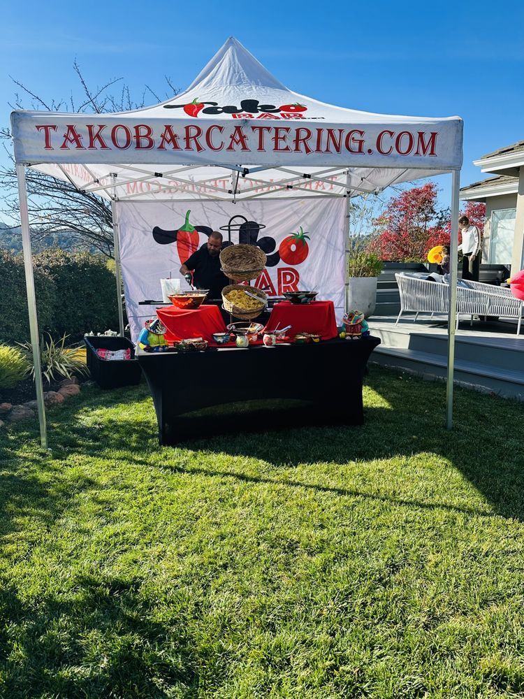 Taco bar catering tent set up on a lawn; the black table is laden with food and toppings.