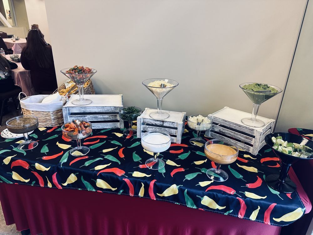 A food display with various dips and toppings in martini glasses, on a table with chili pepper tablecloth.