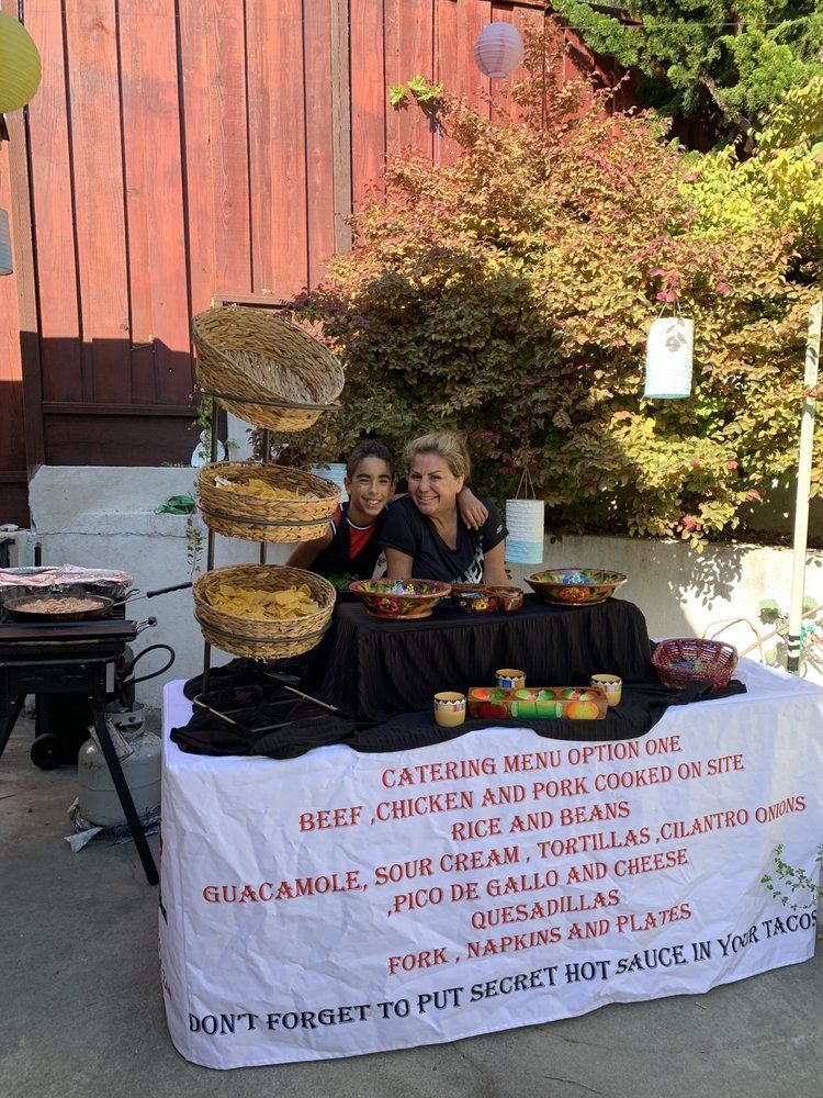 People at a taco catering table, with food displays, offering Mexican cuisine outside.