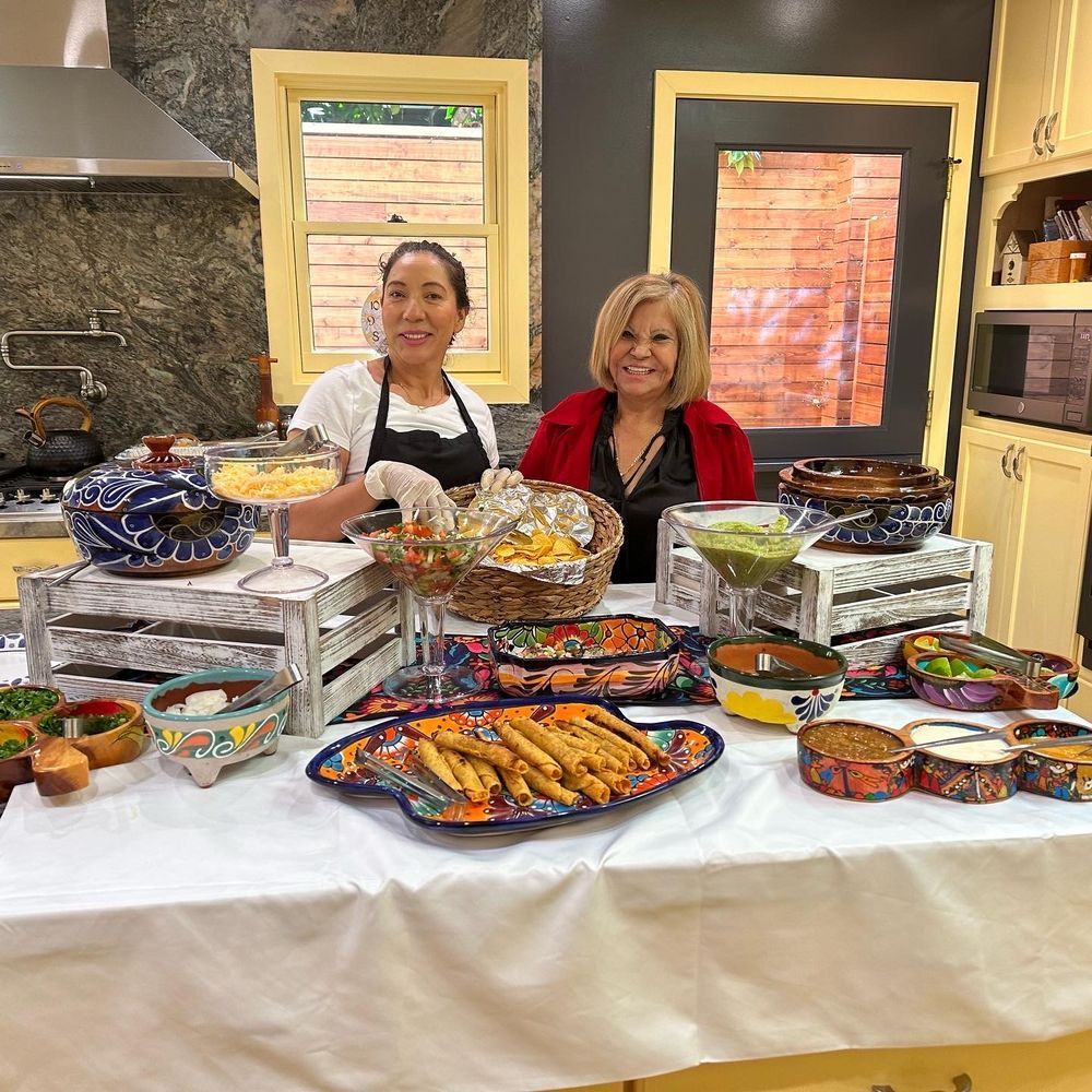 Two women at a table with various Mexican dishes in a kitchen. Smiling, colorful food display.