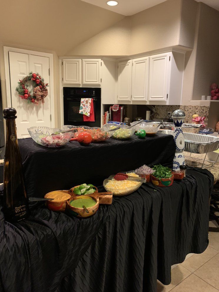 Taco bar setup on a long black-draped table in a kitchen, with various ingredients and bowls.