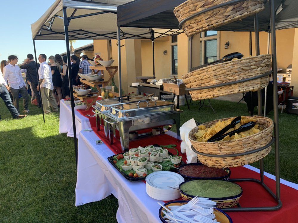 Outdoor buffet with food, serving dishes, and guests under a canopy on a grassy lawn.