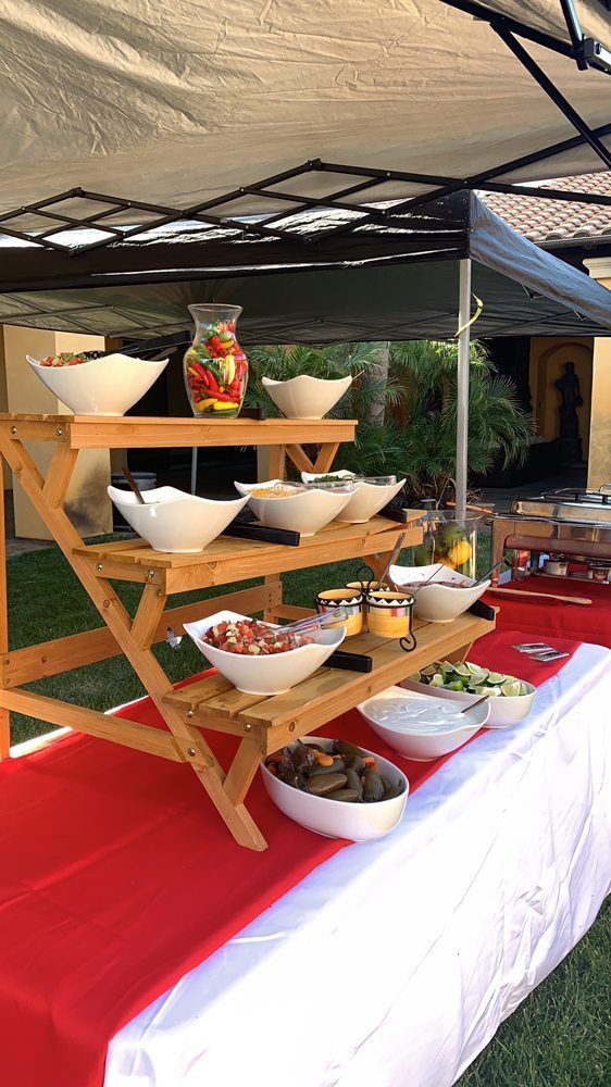 Wooden tiered food display with various dishes, outdoors under a canopy, red table runner.