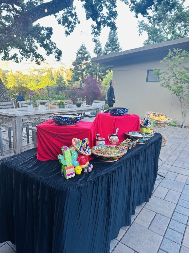 Outdoor buffet table with Mexican decor, red and black tablecloths, and food. Person standing in the background.
