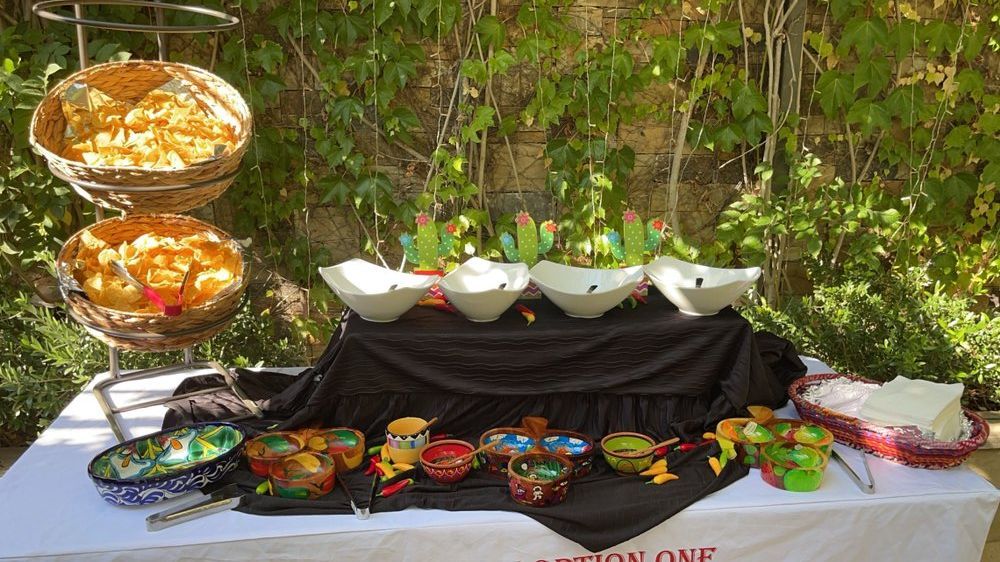 Mexican food buffet with chips, bowls of toppings, and decorative cacti. Outdoor setting.