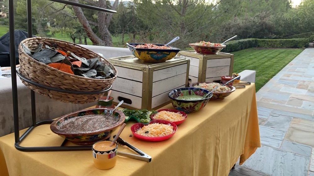 Taco bar on a yellow tablecloth outside: beans, salsa, toppings, and a basket of tortillas.