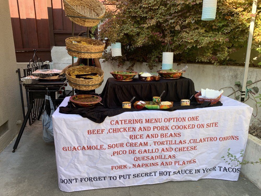 Taco catering setup outdoors with food displayed. Menu sign lists beef, chicken, pork, rice, beans, tortillas and more.