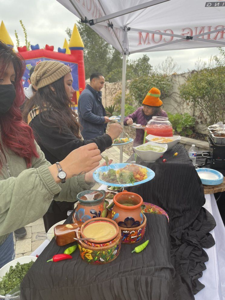 People serving food at a colorful outdoor event, including a person in a orange hat and a bouncy castle in the background.