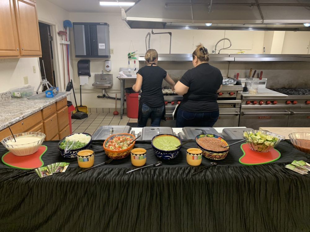 Two women preparing food behind a table of toppings in a kitchen.