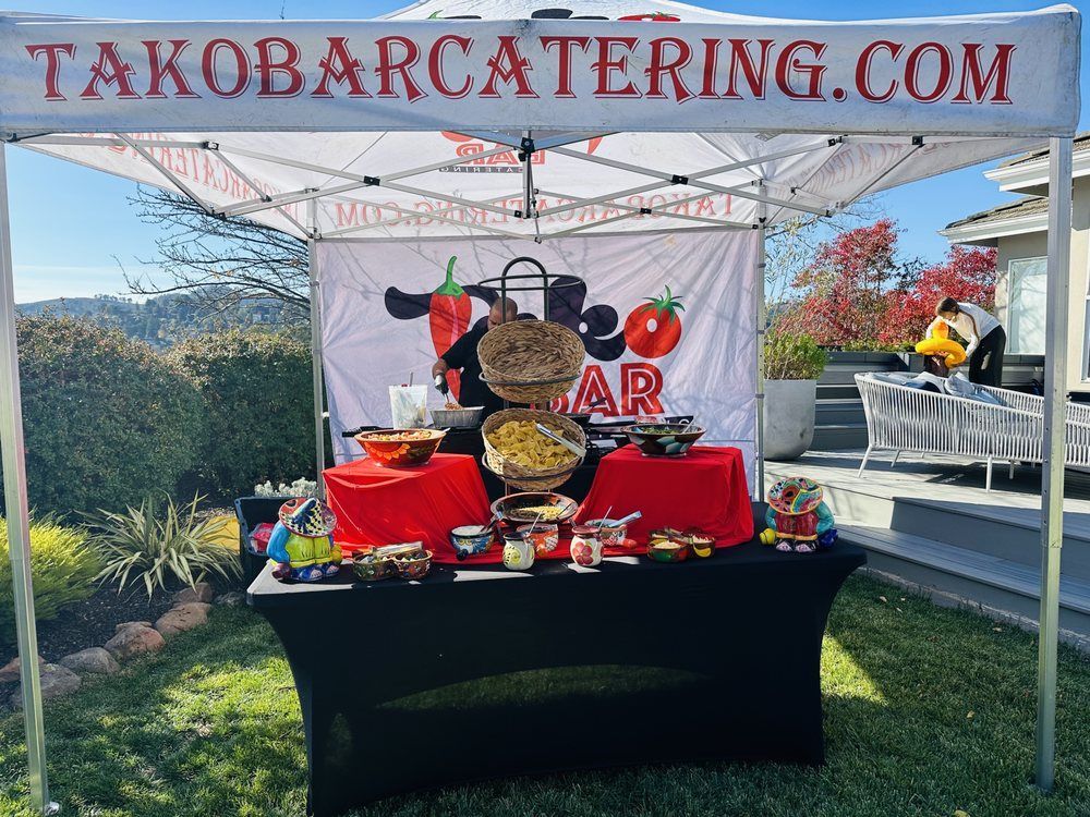 Taco Bar Catering stand set up on a grassy lawn, with food, red table cloths, and a banner with logo.