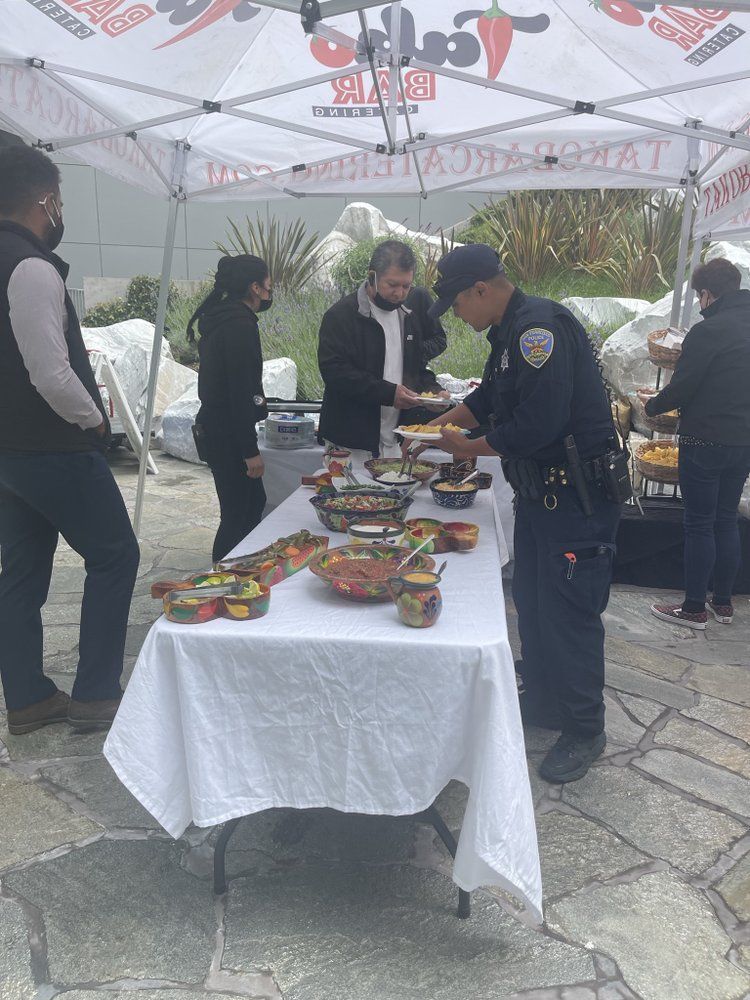 People at outdoor food service table, including a police officer, under a tent.