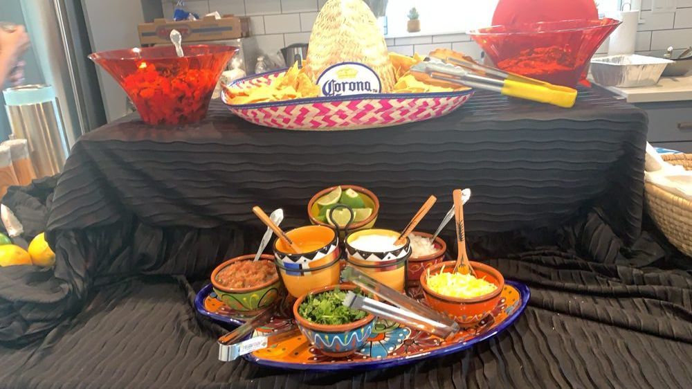Taco bar with chips, toppings in colorful bowls, and Corona sign on a table draped in black fabric.