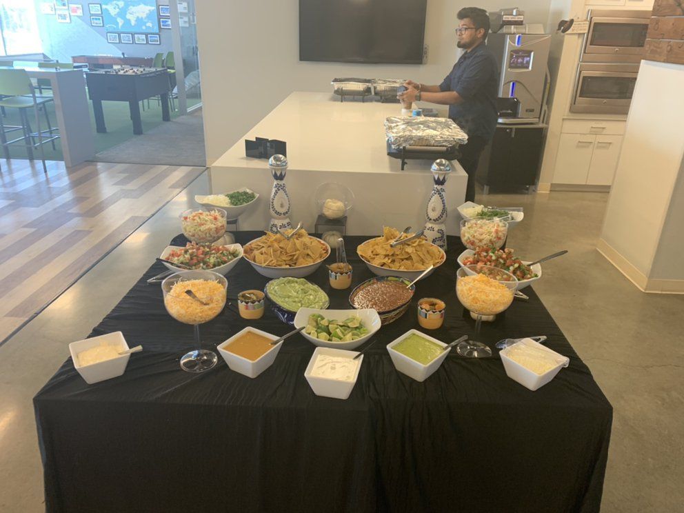 Taco bar with various toppings on a table; a person stands in the background near a kitchen.