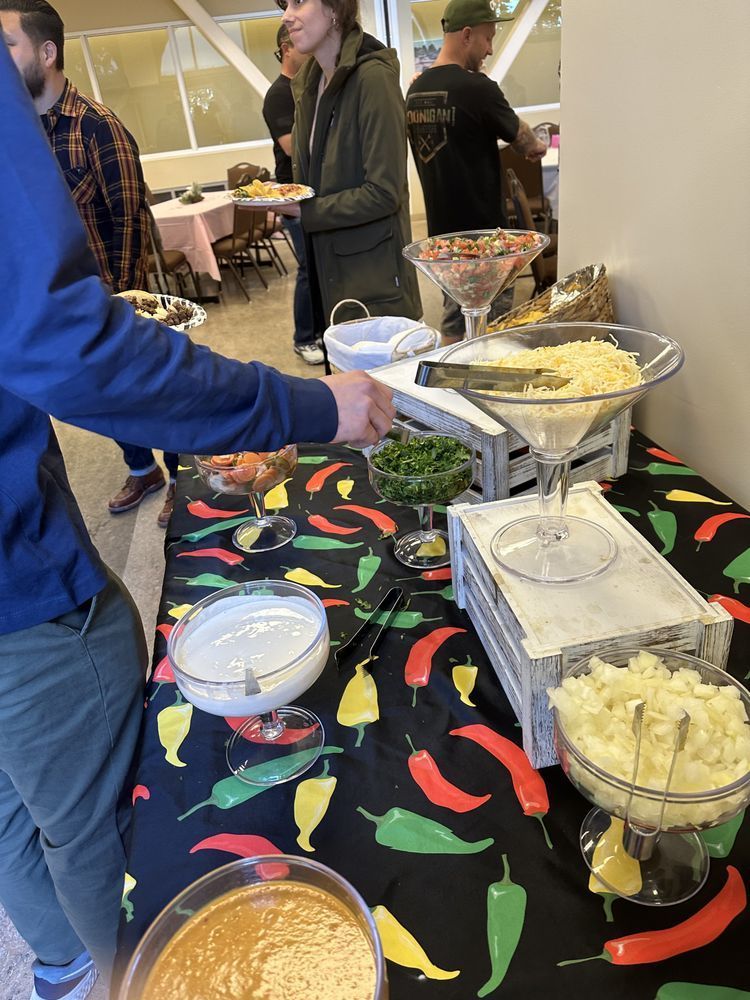 A taco bar with various toppings displayed on a table. People are serving themselves.