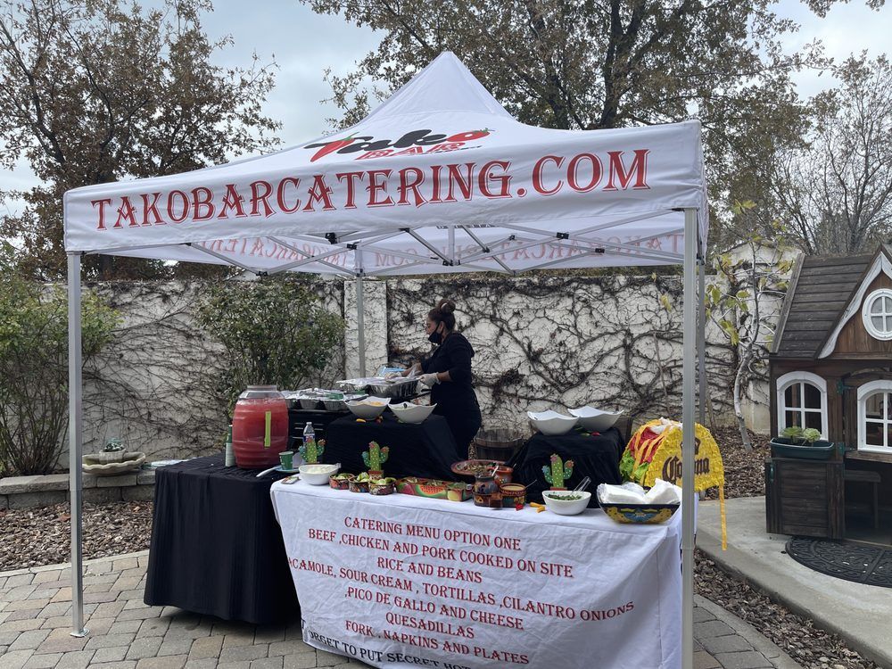 Taco bar catering tent with a person preparing food.  White tent, black table with food. 