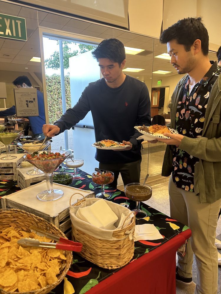 Two men at a buffet table. One serves himself salsa. Chips and various dishes are arranged on the table.