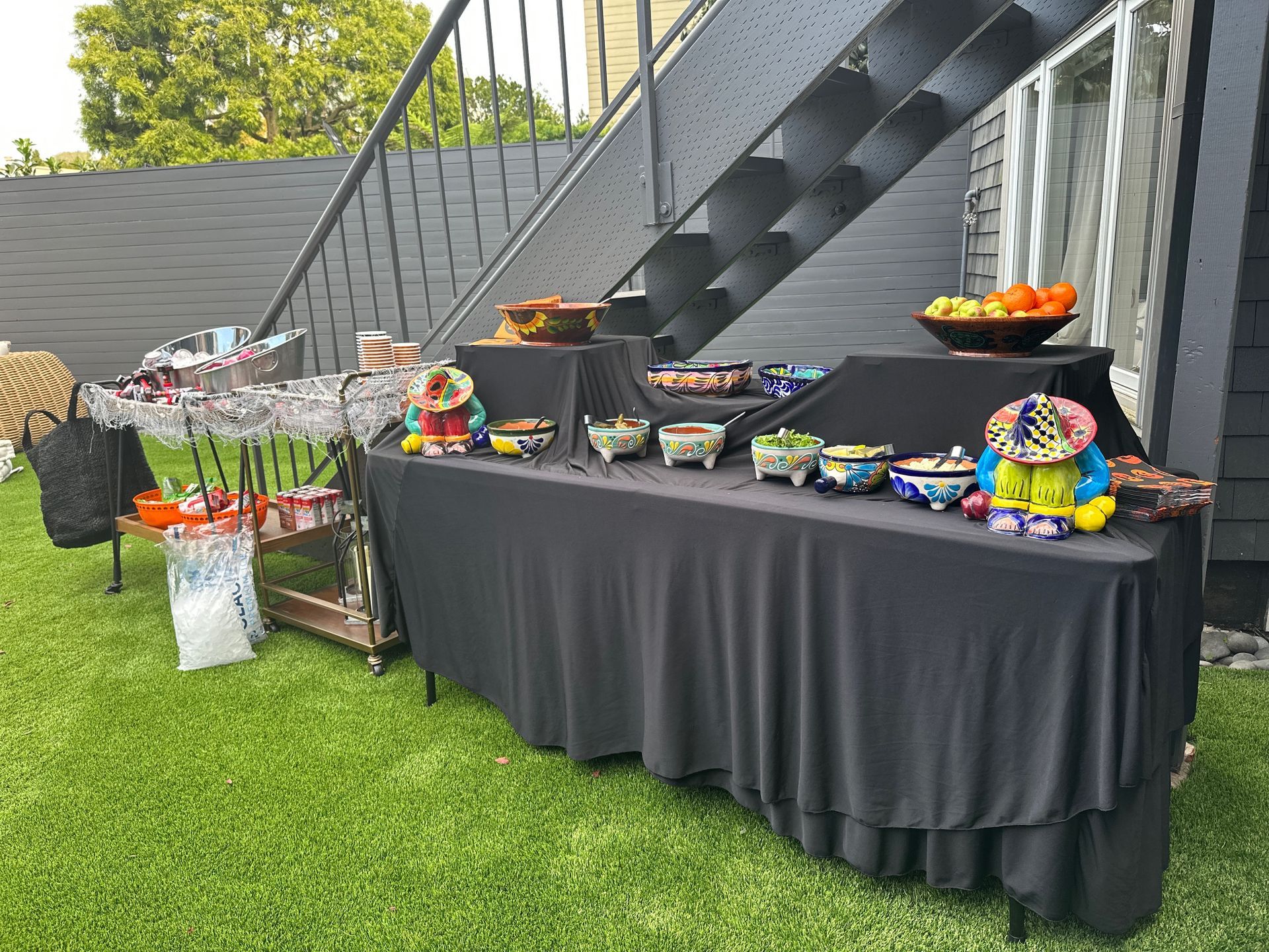 Outdoor party buffet with food in colorful bowls on a black-draped table, next to a stairwell.