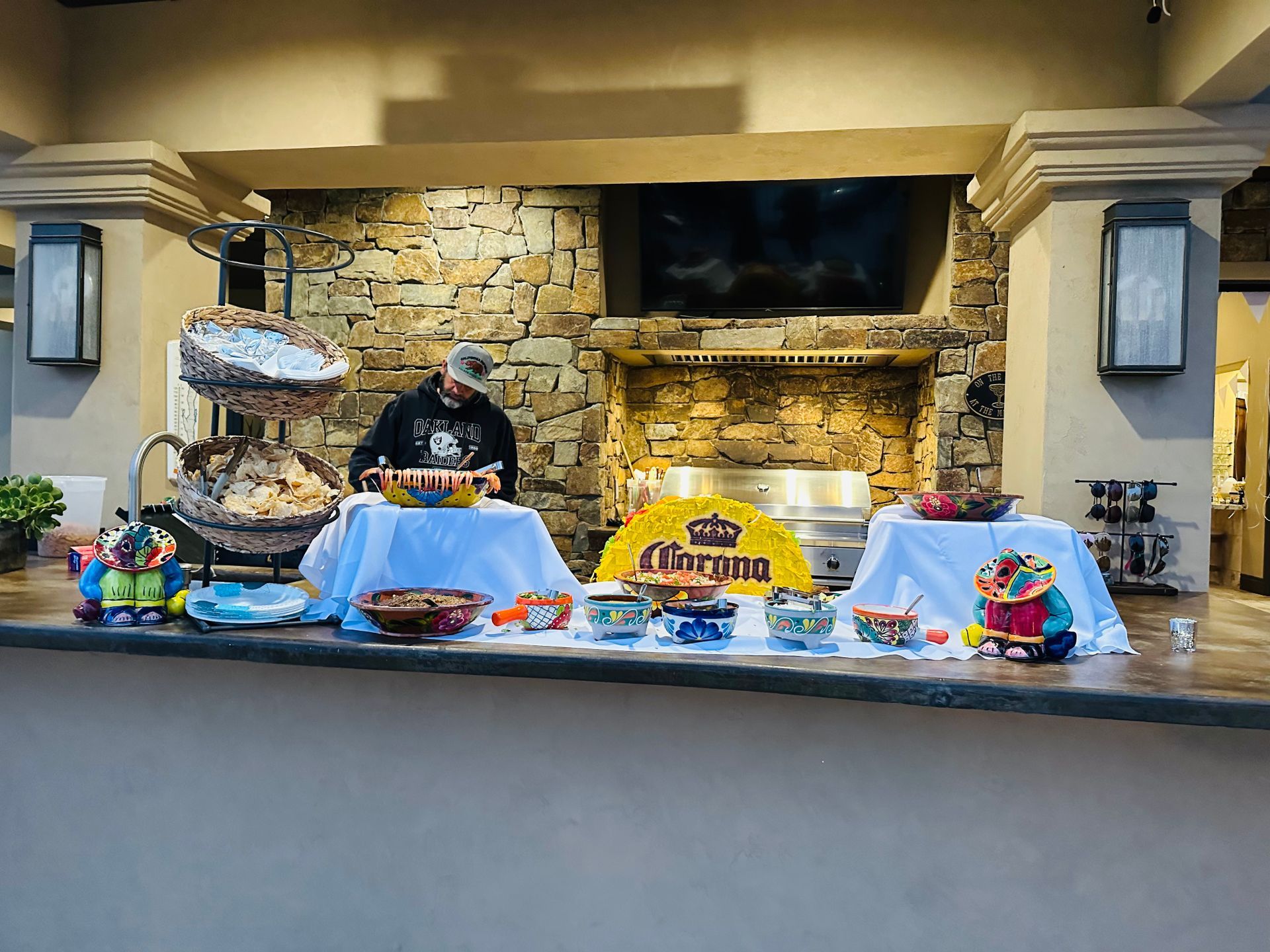 A person sets up a taco bar with colorful bowls, Corona sign, and food, outdoors under a stone wall.
