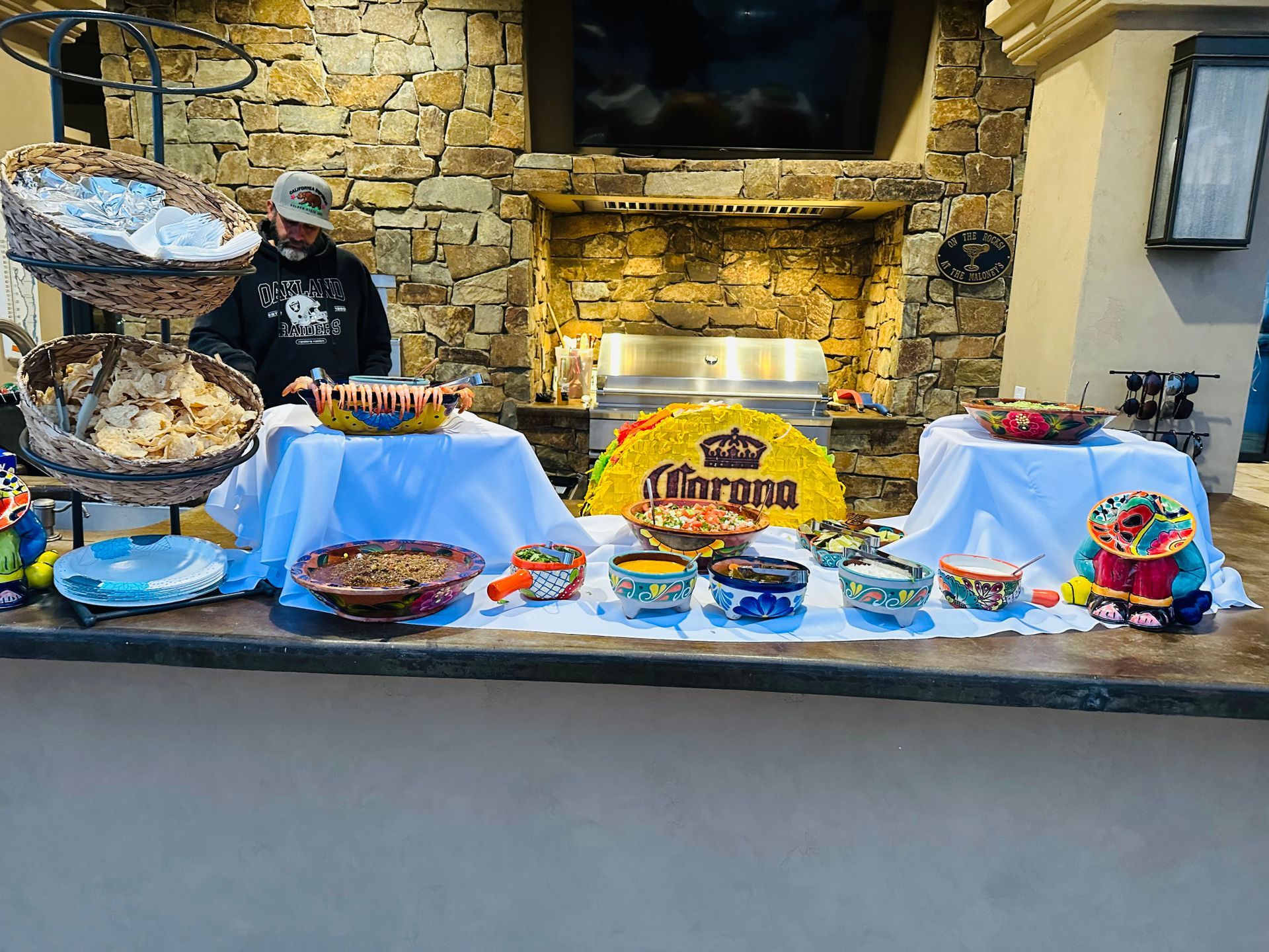 Outdoor bar with food in colorful bowls; man behind it. Corona sign in the middle.