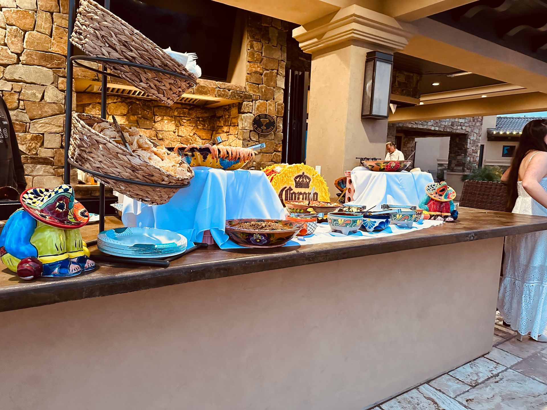 Buffet with pottery, food, and Corona banner in a stone-walled outdoor area. Woman in white dress stands nearby.
