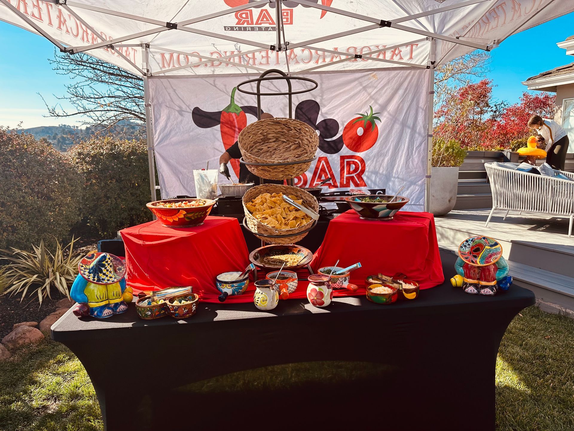 Taco bar setup outdoors with red tablecloths, bowls of food, and a sign.