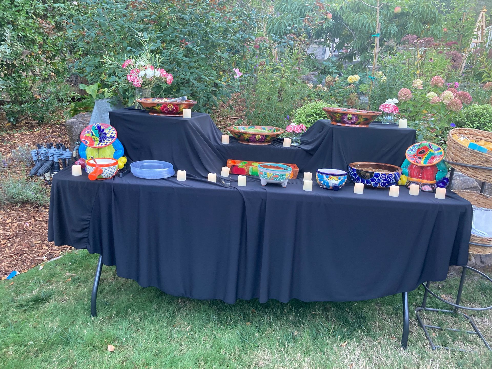 A black table with colorful bowls, candles, and decor in a garden setting.