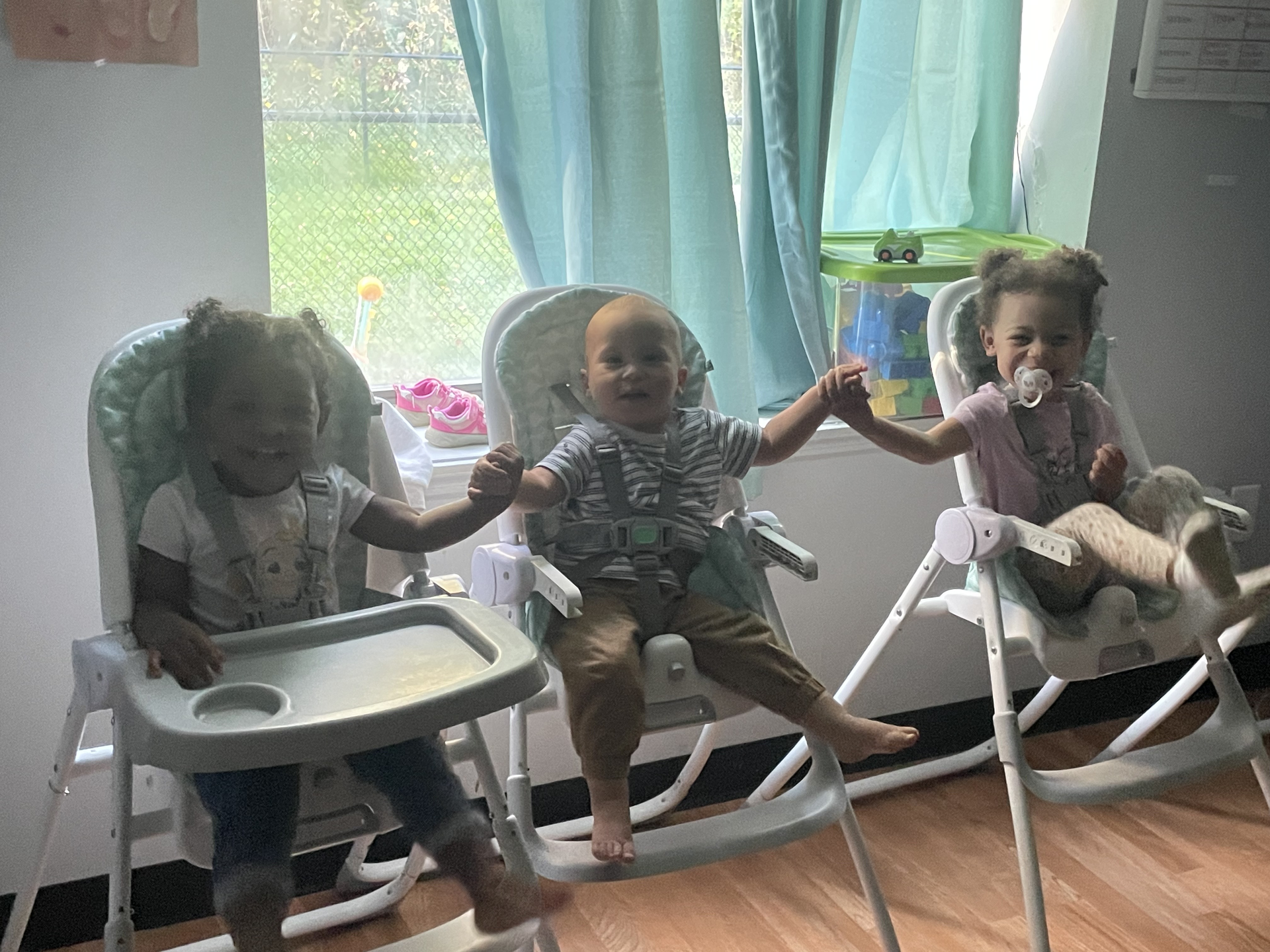 Three babies are sitting in high chairs holding hands.
