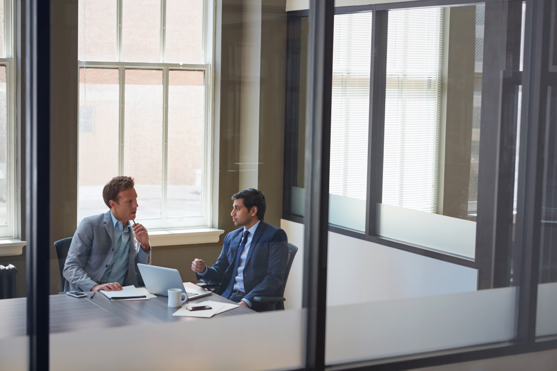 Two professionals in suits talking at a table in a glass-walled office meeting room