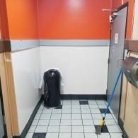 Narrow tiled hallway with orange and white walls, a trash can, and a mop leaning by a door.