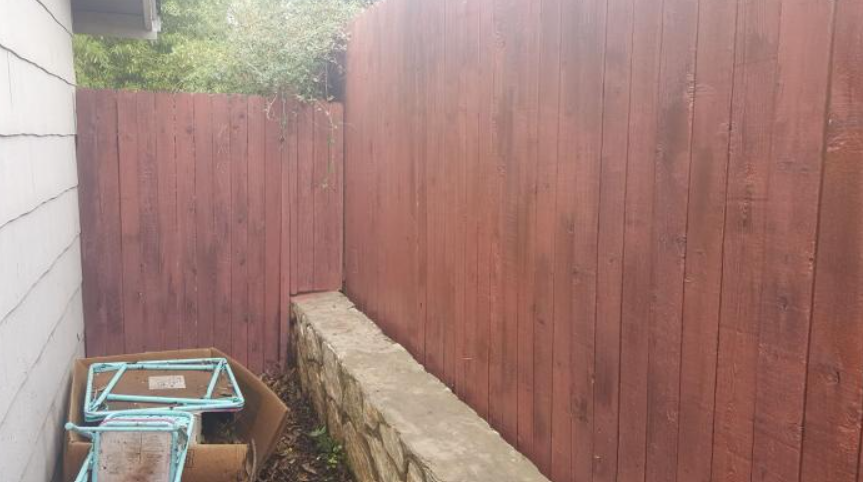 Narrow backyard with a tall red wooden fence, stone retaining wall, and a rusty wheelbarrow in the corner.