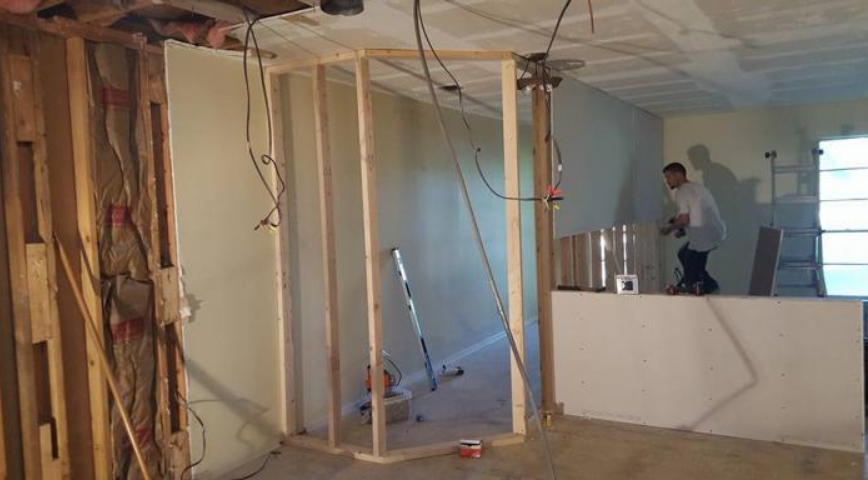 Interior under renovation with exposed framing, hanging wires, and a worker standing on a raised platform.