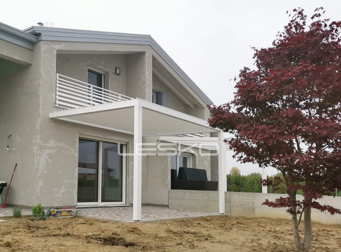 Una moderna casa beige a due piani con un pergolato bianco che copre un patio, adiacente a un albero dalle foglie rosse.