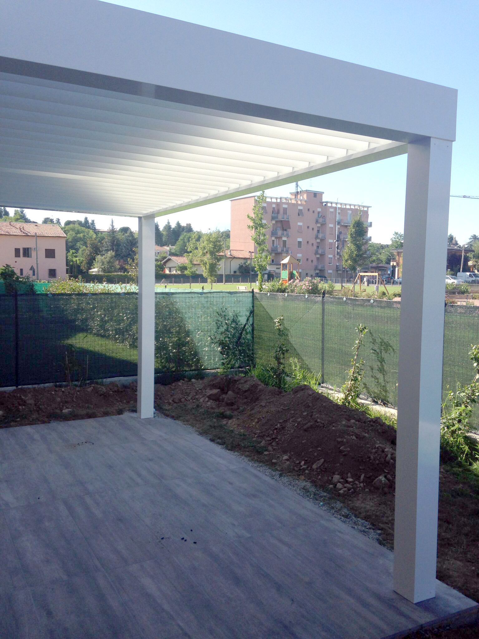 Un pergolato bianco con lamelle orientabili sopra un patio, con un'area adibita a giardino e una recinzione in rete verde sullo sfondo.