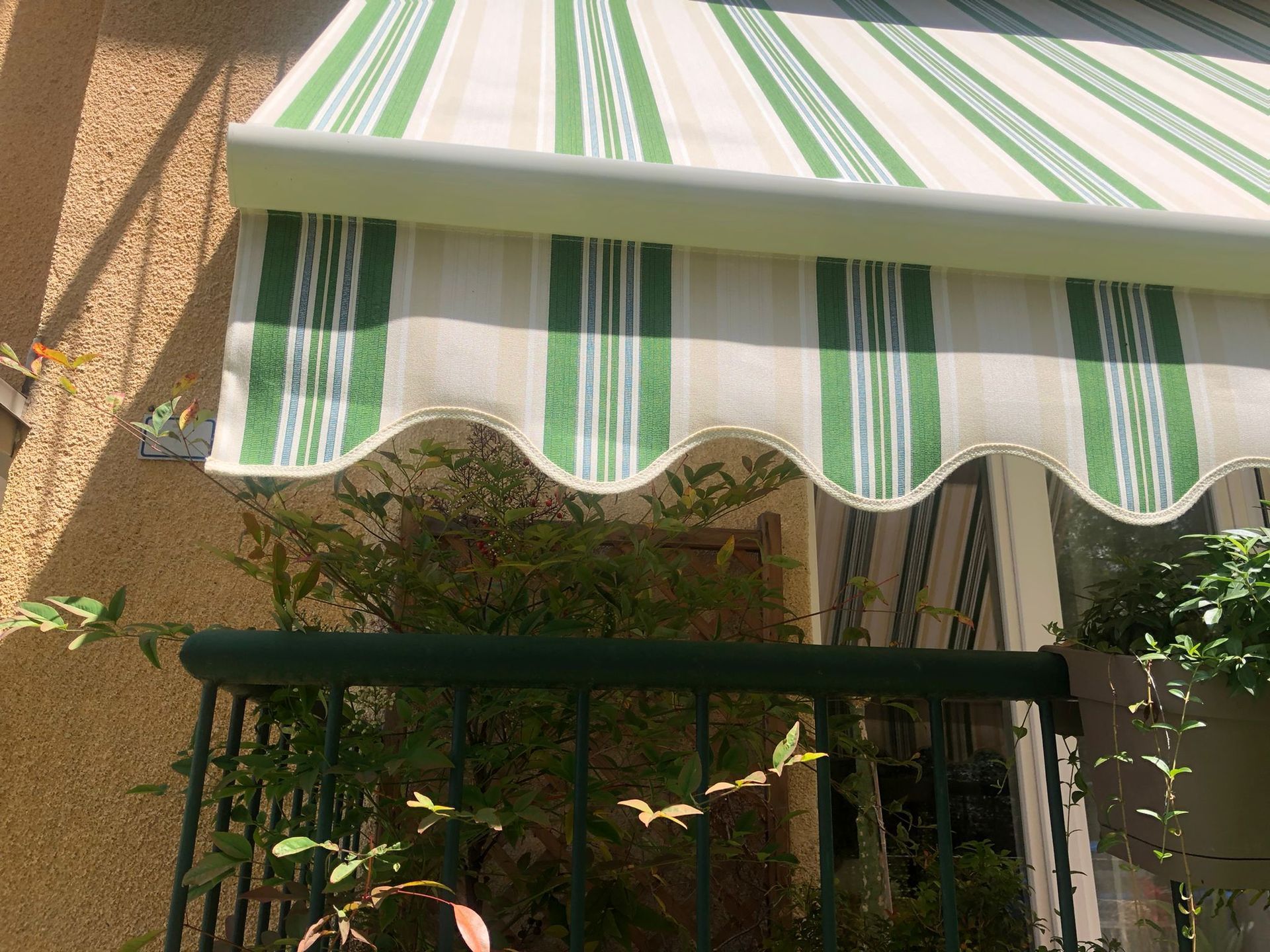 Una tenda da sole in tessuto a righe verdi e bianche sopra una ringhiera scura ricoperta di vegetazione in una giornata di sole.