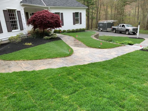 Suburban front yard with a curved stone walkway, green lawn, red shrub, and a parked pickup truck by the house