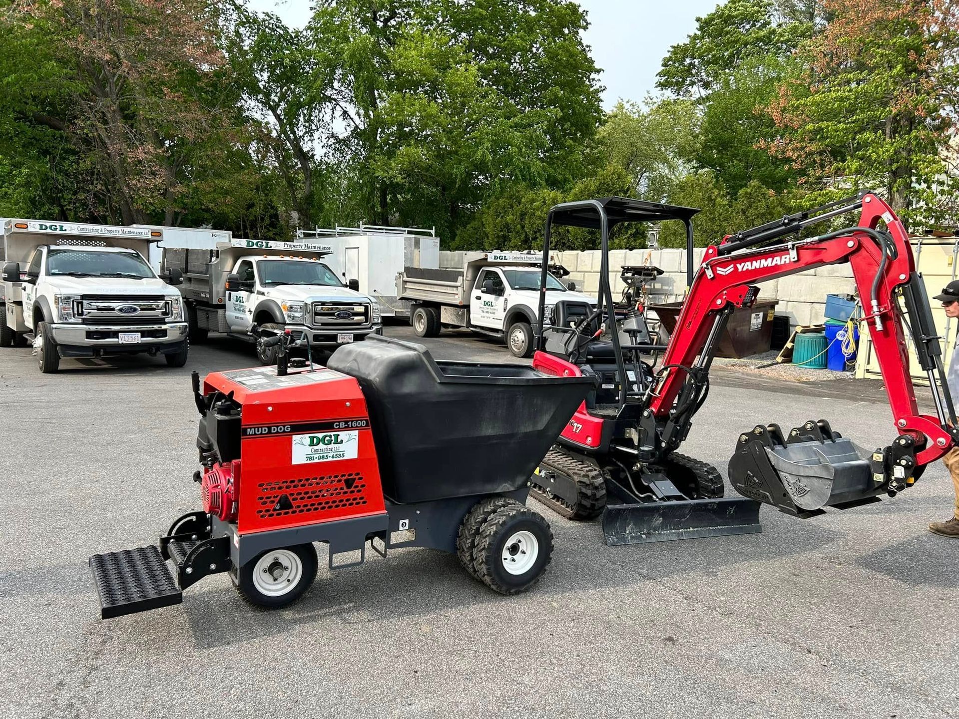 Red-and-black mini excavator with loader bucket in a gravel lot, trucks parked behind it