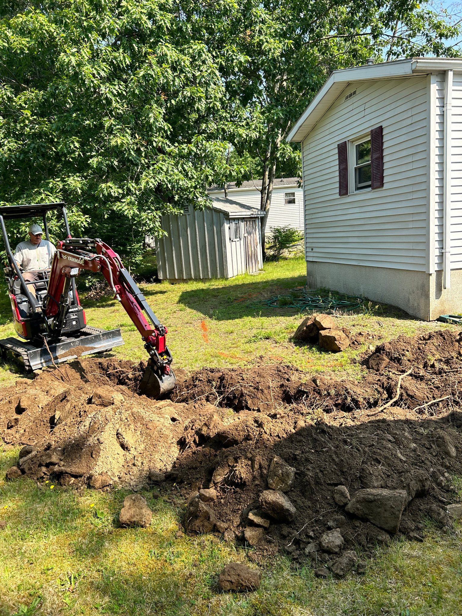 Backhoe digging a trench beside a white house in a grassy yard on a sunny day