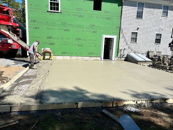 Workers pouring a new concrete slab beside a green building and a stone wall