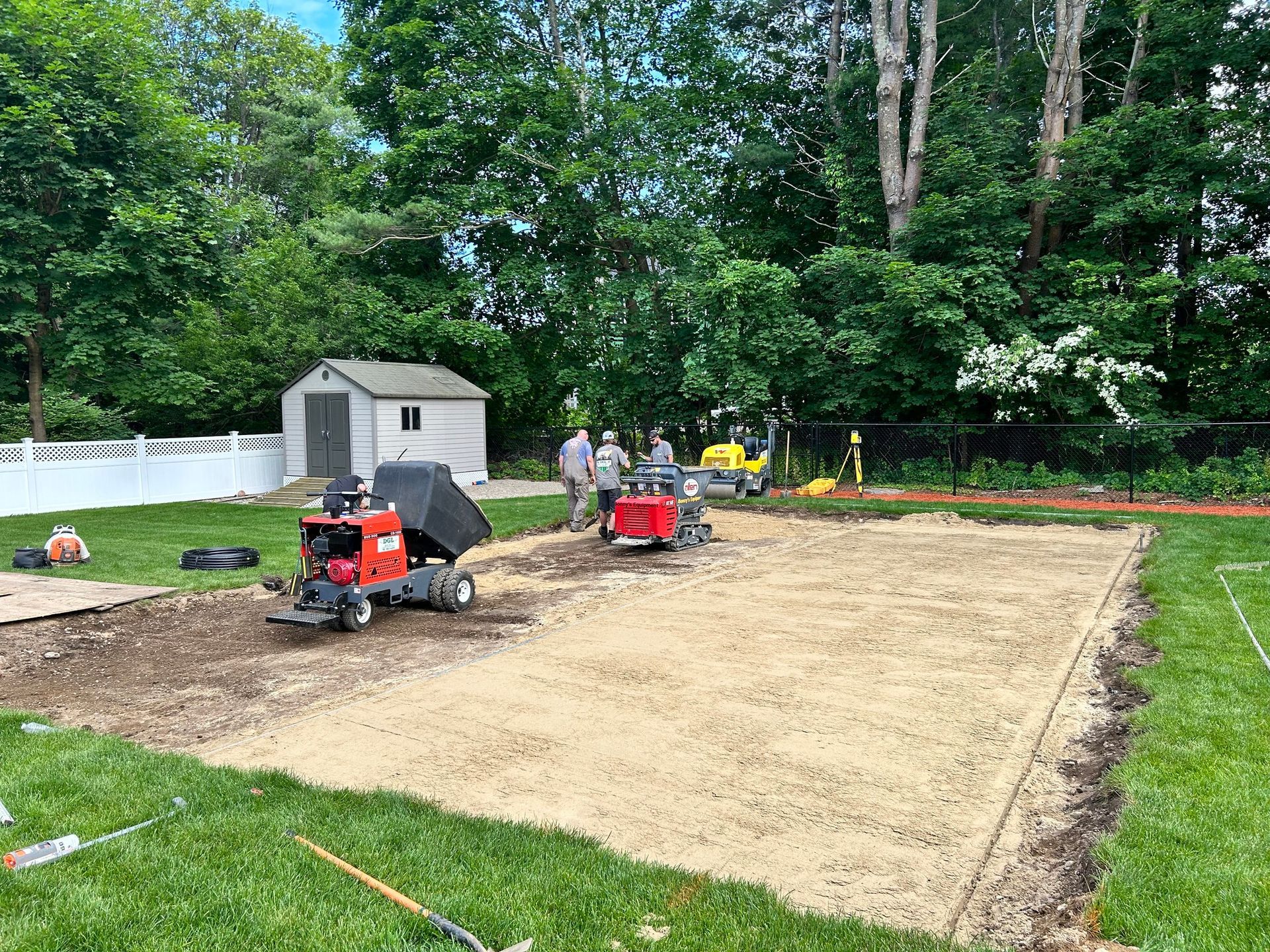 Two red compact tractors level soil in a backyard beside a shed and trees.