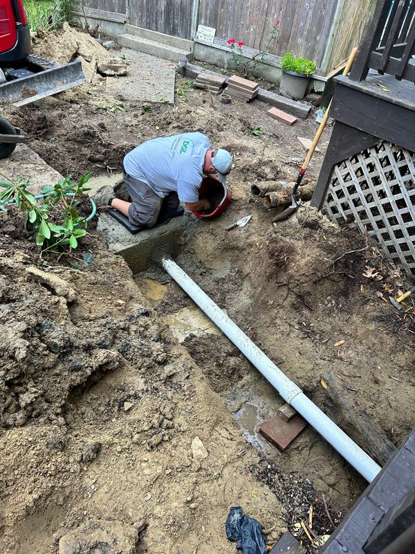 Person kneeling in a muddy backyard trench, working near a white pipe and wooden fence.